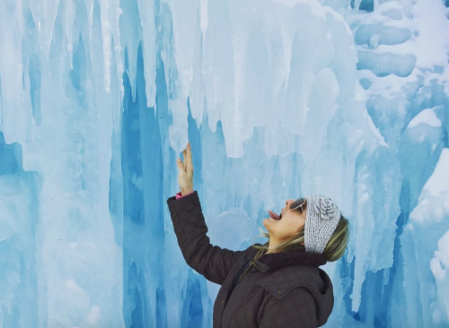 A woman in a winter coat and sunglasses playfully reaching towards icicles in a blue ice formation.