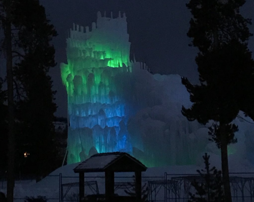 A towering ice structure illuminated with green and blue lights, surrounded by trees at night.