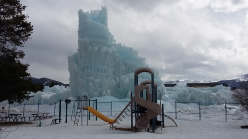 A large ice structure towers over a playground with a slide, surrounded by snow and cloudy skies.