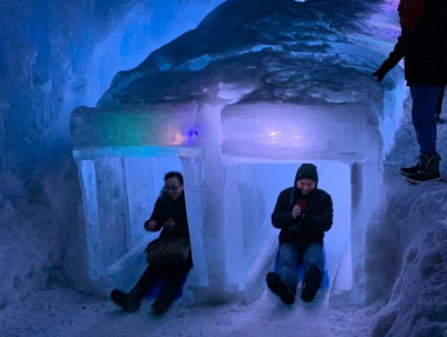 Two people sitting in ice structures, illuminated by colorful lights, inside a snowy cave.