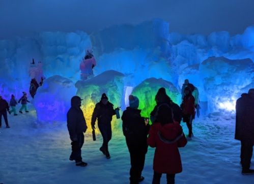 People explore a colorful ice sculpture display at night, illuminated by vibrant blue, green, and purple lights.