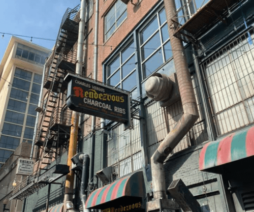 Sign for "Charles Vergos' Rendezvous Charcoal Ribs" on a brick building with fire escapes and awnings.