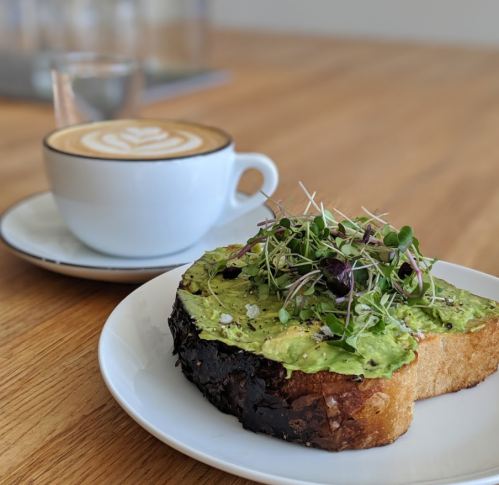 A slice of avocado toast topped with microgreens, next to a latte with a leaf design in a white cup on a wooden table.