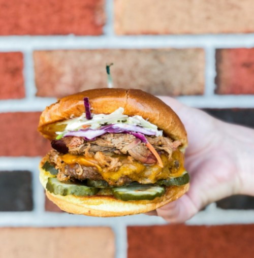 A hand holding a loaded burger with pulled pork, cheese, pickles, and coleslaw against a brick wall background.