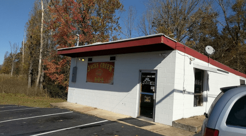 A small, white building with a red roof and sign reading "Jake's Grey" surrounded by trees and a clear blue sky.
