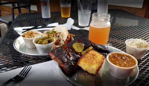 A plate of barbecue ribs with sides of cornbread, baked beans, and coleslaw, alongside drinks on a table.