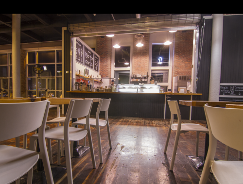 Interior of a café with empty white chairs, a counter displaying menu items, and large windows letting in light.