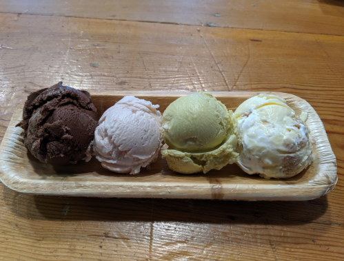 A wooden tray holds four scoops of ice cream: chocolate, strawberry, pistachio, and vanilla with nuts.