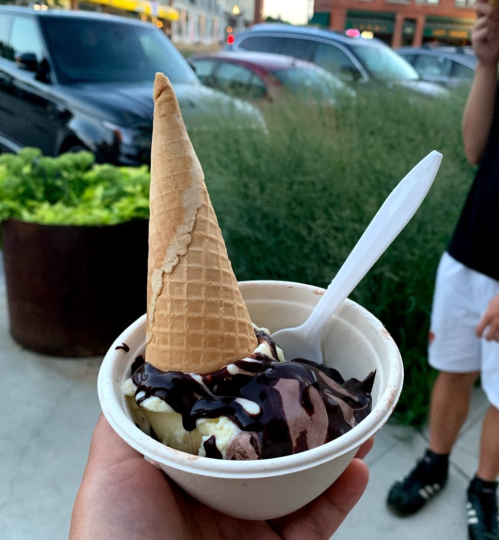 A hand holding a bowl of ice cream topped with chocolate sauce and a cone, with cars and greenery in the background.