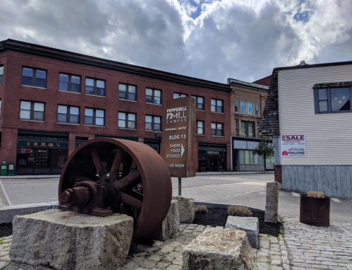 A large rusted wheel on stone blocks in front of a building with a sign for Pepperell Mill Campus and shops.