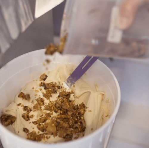 A hand pours granola over creamy ice cream in a container, with a purple spatula resting inside.