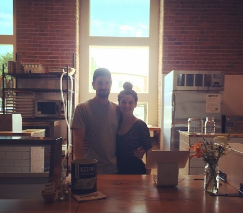 A couple stands together in a bright, rustic café with brick walls and a wooden counter.