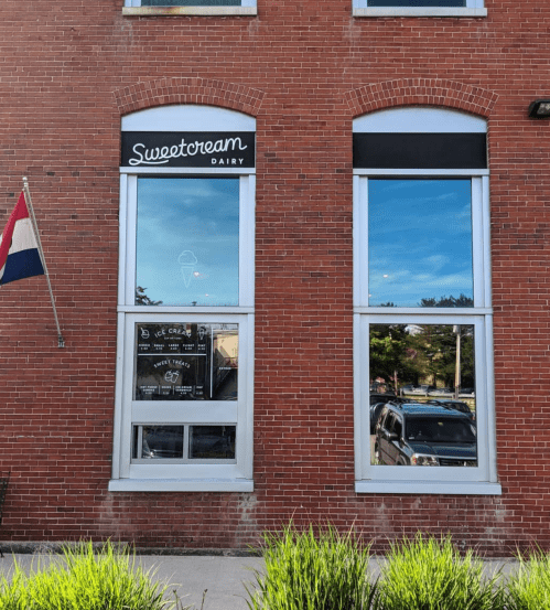 Exterior of Sweetcream Dairy with large windows, a sign, and a flag in front of a brick building.