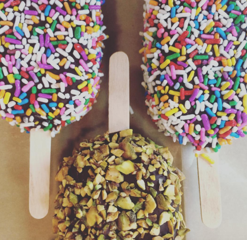 Three ice cream bars: two covered in colorful sprinkles and one coated in chopped pistachios, all on wooden sticks.