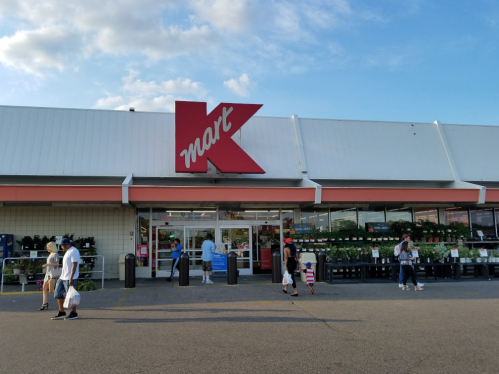 Exterior of a Kmart store with shoppers entering and exiting, surrounded by greenery and a clear blue sky.