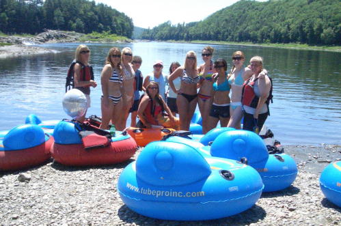 A group of people in swimsuits poses by the river with colorful inflatable tubes on a sunny day.