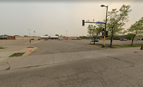 Empty parking lot at an intersection, with traffic lights and nearby retail buildings under a hazy sky.