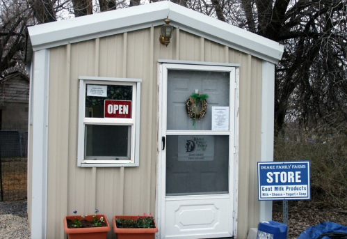 A small metal building with a sign reading "Open" and a store sign for goat milk products, surrounded by potted plants.