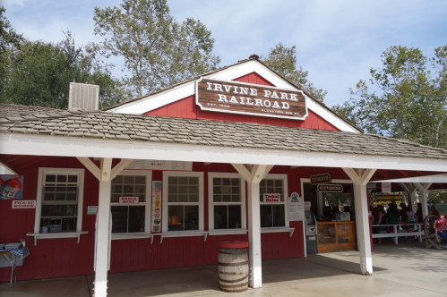 Red and white building with a sign reading "Irvine Park Railroad," surrounded by trees and a clear sky.