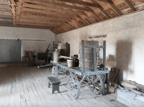 An old wooden cart with barrels and milk cans inside a rustic, empty room with wooden floors and a sloped ceiling.
