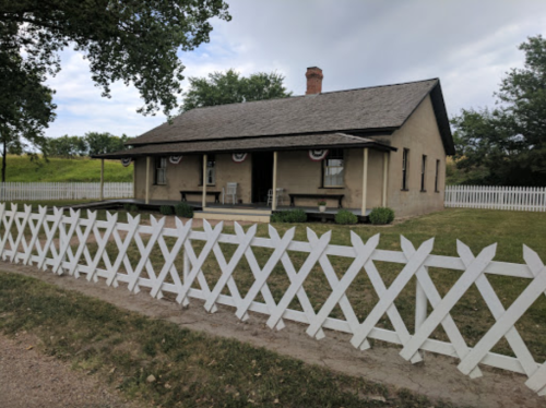 A historic house with a porch, surrounded by a white picket fence and greenery under a cloudy sky.