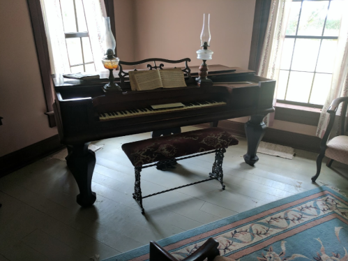 A vintage piano with sheet music, a decorative bench, and oil lamps in a sunlit room with large windows.