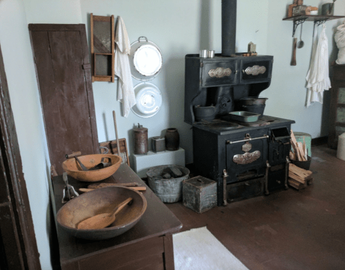 A vintage kitchen with a black stove, wooden bowls, utensils, and various kitchen items on shelves and tables.