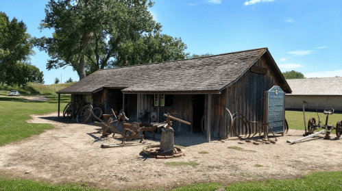 A rustic wooden barn surrounded by vintage farming equipment on a grassy area under a clear blue sky.