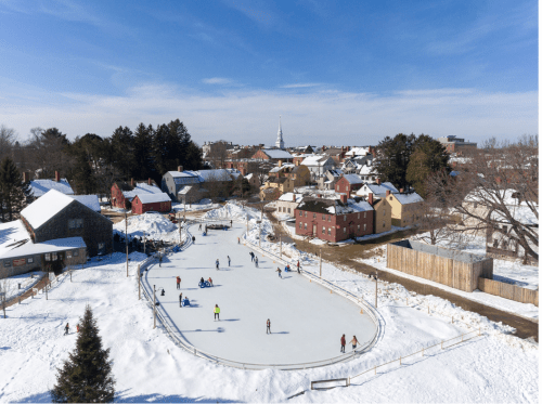 Aerial view of an outdoor ice skating rink surrounded by snow-covered houses and trees under a clear blue sky.