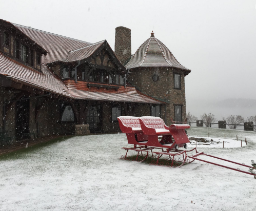 A snowy scene featuring a large stone house and a red sleigh on a white lawn. Snowflakes are falling gently.