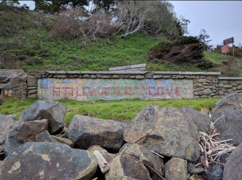 Colorful "Stillwater Cove" sign on a stone wall, surrounded by grass and rocks. Trees in the background.