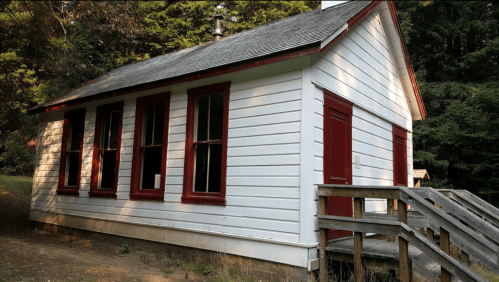 A small, white wooden building with red trim and steps, surrounded by trees in a natural setting.