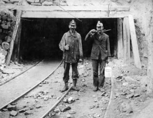 Two miners stand outside a mine entrance, holding tools and buckets, with railway tracks visible in the foreground.