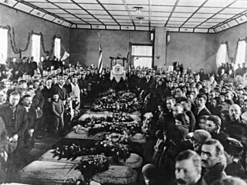 A large crowd gathers in a room filled with flowers and caskets, mourning a significant event or loss.