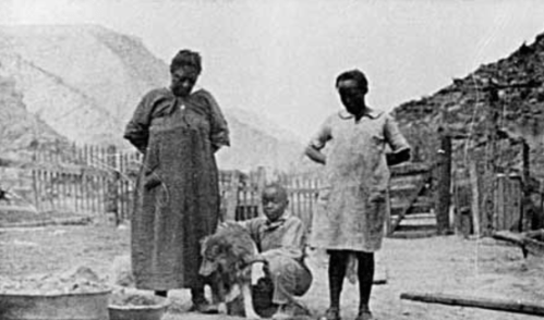 Two women and a child stand outdoors with a dog, surrounded by a rustic setting and wooden structures.