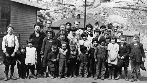 A historical black-and-white photo of a group of children standing together outdoors, with buildings and rocky terrain in the background.