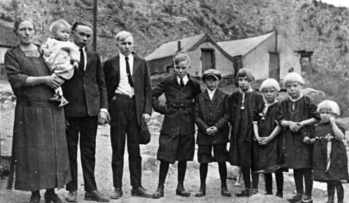 A historical black-and-white photo of a group of children and adults standing together outdoors, with houses in the background.