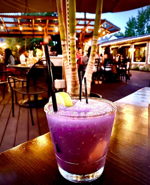 A vibrant purple cocktail with lime, served in a glass with a salted rim, set against a lively outdoor dining scene.
