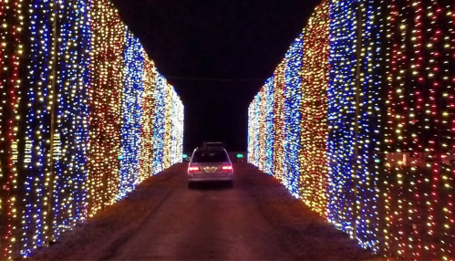 A car drives through a tunnel of colorful holiday lights on either side, creating a festive atmosphere at night.