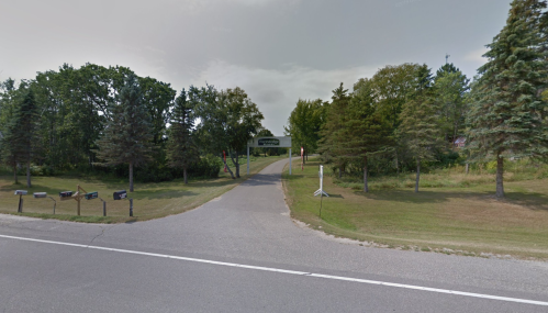 A tree-lined road leads to a driveway, with mailboxes on the left and a sign in the distance.