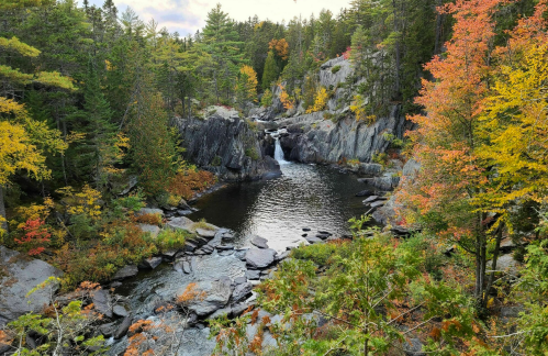 A serene waterfall cascades into a calm pool, surrounded by vibrant autumn foliage and rocky cliffs.