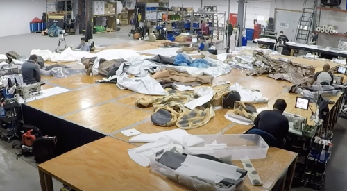 A busy workshop with workers sewing large fabric pieces on tables, surrounded by various tools and materials.