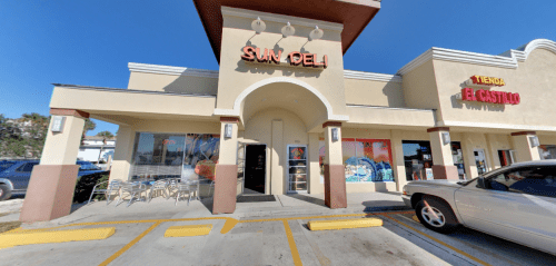 Exterior of a deli named "Sun Deli" with colorful signage, outdoor seating, and a nearby store called "Tienda El Castillo."