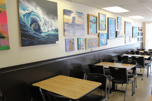 A cozy café interior featuring wooden tables and a wall adorned with various colorful paintings.
