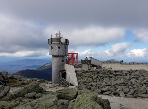 A stone tower with antennas stands on rocky terrain, surrounded by clouds and mountains in the background.