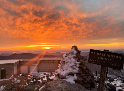 Sunset over Mt. Washington summit, with a stone cairn and sign, snow on the ground, and vibrant orange clouds.