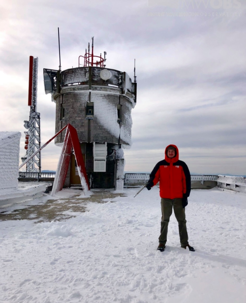 A person in a red jacket stands on a snowy rooftop near a weather tower, with overcast skies in the background.