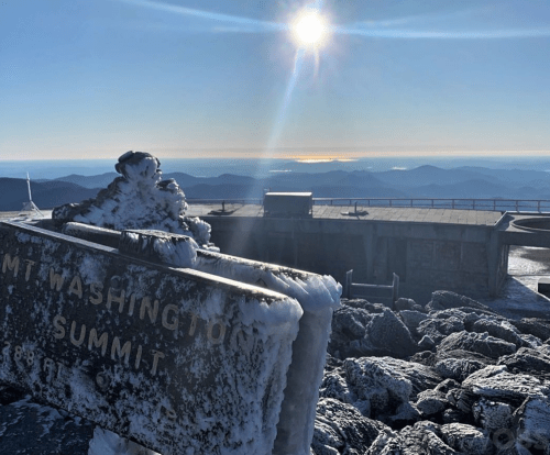 Snow-covered summit sign at Mount Washington, with a bright sun shining over distant mountains and a clear blue sky.