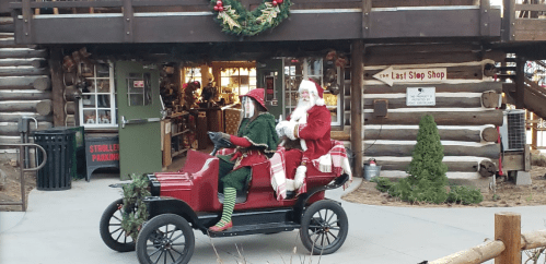 Santa and an elf ride in a vintage car, decorated for the holidays, in front of a log cabin shop.