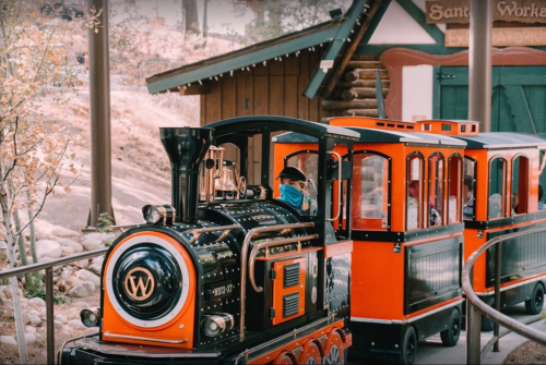 A colorful miniature train with a black engine and orange cars, parked near a wooden building in a scenic outdoor setting.